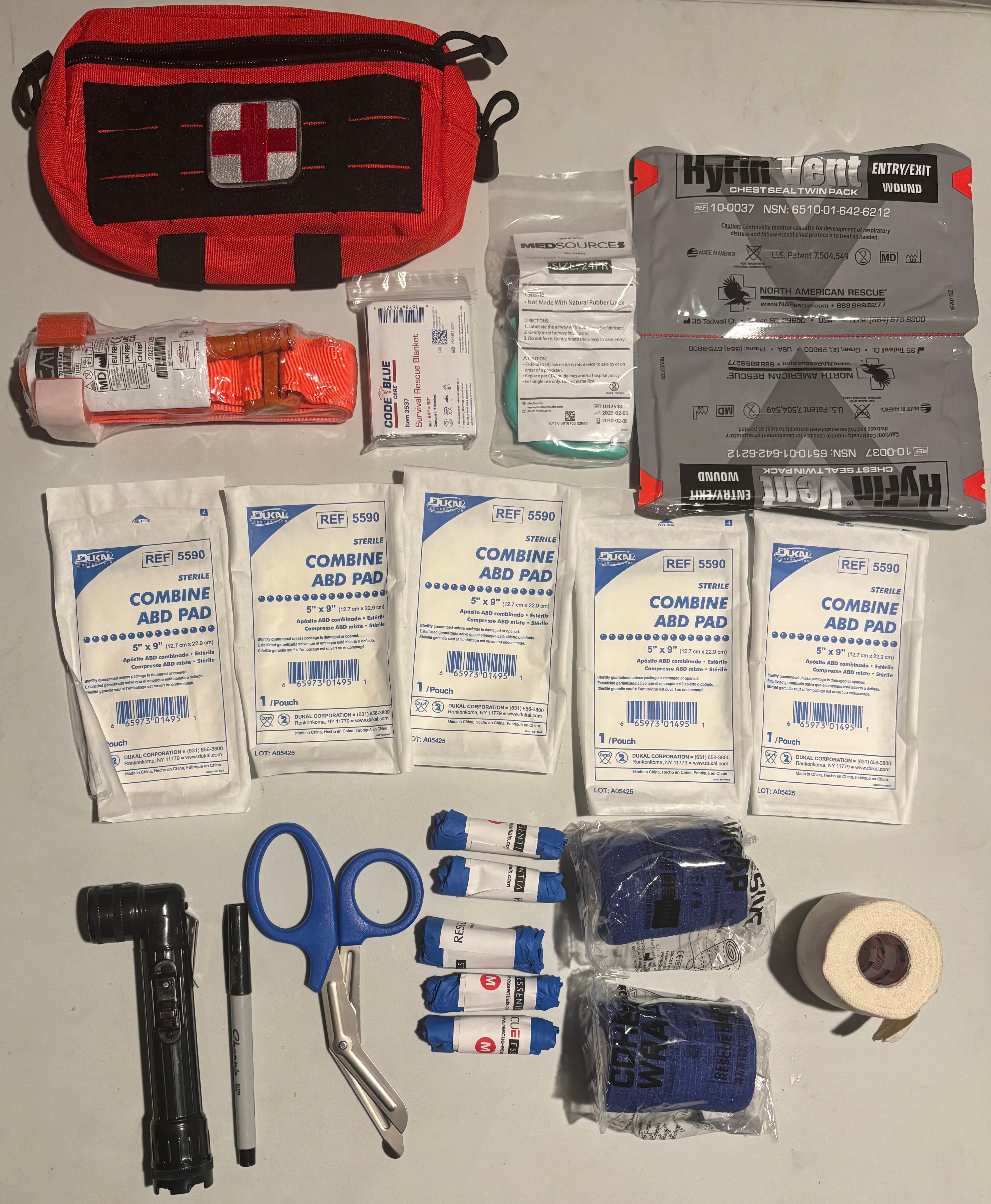 Kit includes pouch with a white cross, tourniquet, Mylar blanket,  Nasopharyngeal airway, pair of vented chest seals, 5- 5 x 9 dressings, flashlight, permanent marker, trauma shears, 5 pairs of nitrile gloves, 2 rolls self adhering bandages, 1 roll 2 inch cloth tape